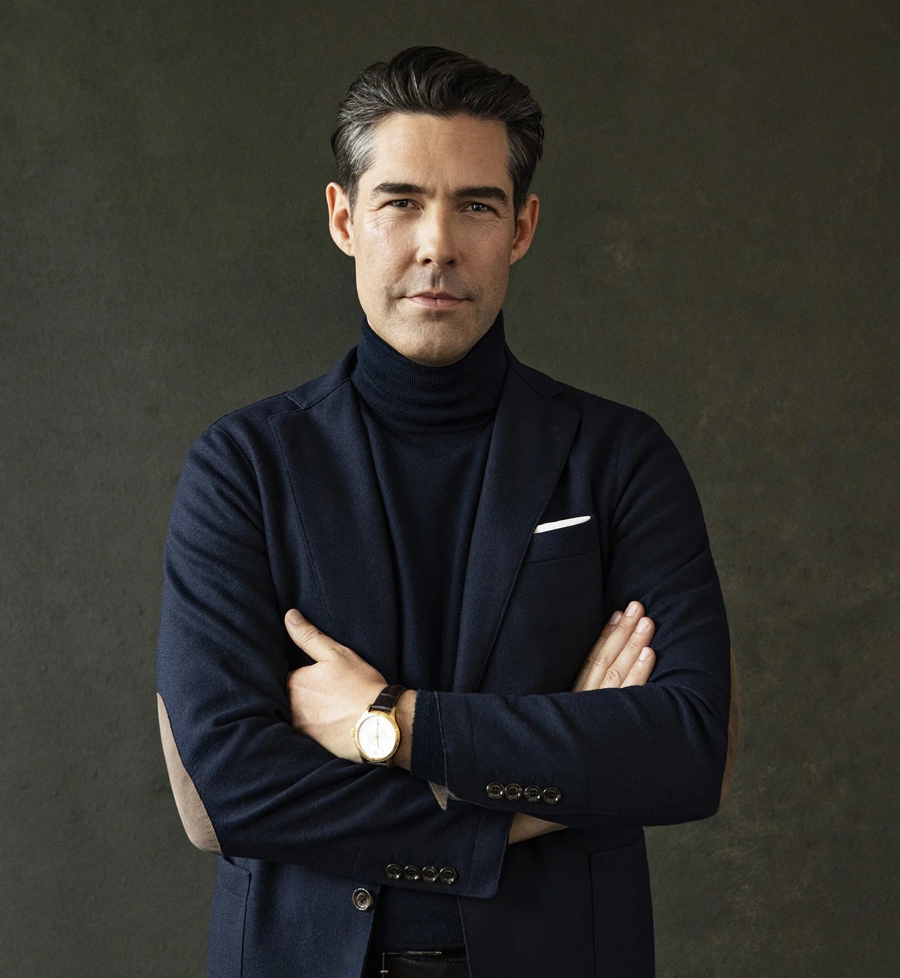 Michael Beglinger trägt einen dunkelblauen Anzug mit schwarzen Turtleneck, seine Arme sind verschränkt, er trägt eine Armbanduhr.